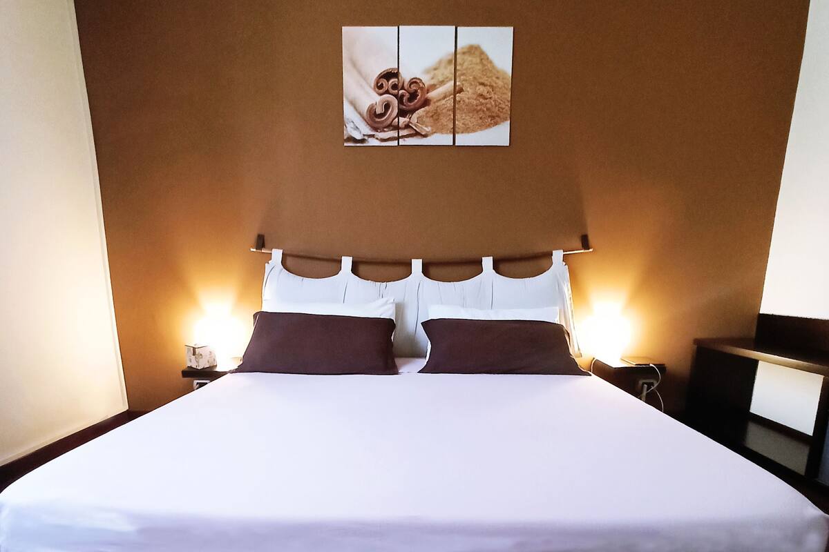 A neatly made bed with white linens and two brown pillows, a wall-mounted artwork above it, and a nightstand with a lamp to the right.