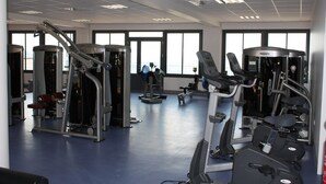 A gym with various exercise machines, large windows, and a blue floor. - Village de gîtes les hauts du Gevaudan (Montrodat)