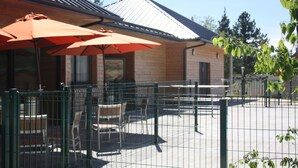 An outdoor patio area with tables and chairs, enclosed by a green fence, and featuring orange umbrellas. - Village de gîtes les hauts du Gevaudan (Montrodat)