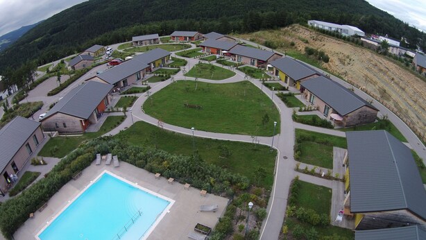 A resort with multiple chalet-style buildings arranged around a central lawn, a swimming pool, and a hilly backdrop. - Village de gîtes les hauts du Gevaudan (Montrodat)