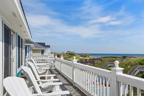 Un balcon avec des chaises blanches, une terrasse en bois et une vue sur la plage et les cabanes de plage.