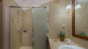 A bathroom with a glass shower enclosure, a sink, a mirror, and a small shelf with toiletries. - Rodia House (Rhodes)
