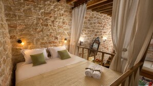 A bedroom with a stone wall, a bed with white bedding and green pillows, a wooden headboard, and a small table with towels. - Rodia House (Rhodes)
