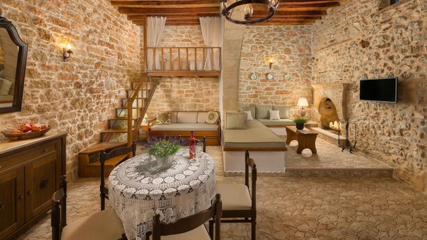 A rustic stone-walled room with a wooden ceiling, a round table with a lace tablecloth, a sofa, a TV mounted on the wall, and a staircase leading to a loft area. - Rodia House (Rhodes)