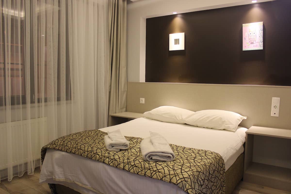 A hotel room with a bed, two bedside tables, a window with sheer curtains, and a wall-mounted light fixture.