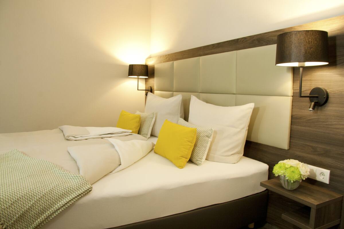 A neatly made bed with white and yellow pillows, a green throw blanket, and a small vase with flowers on a bedside table.