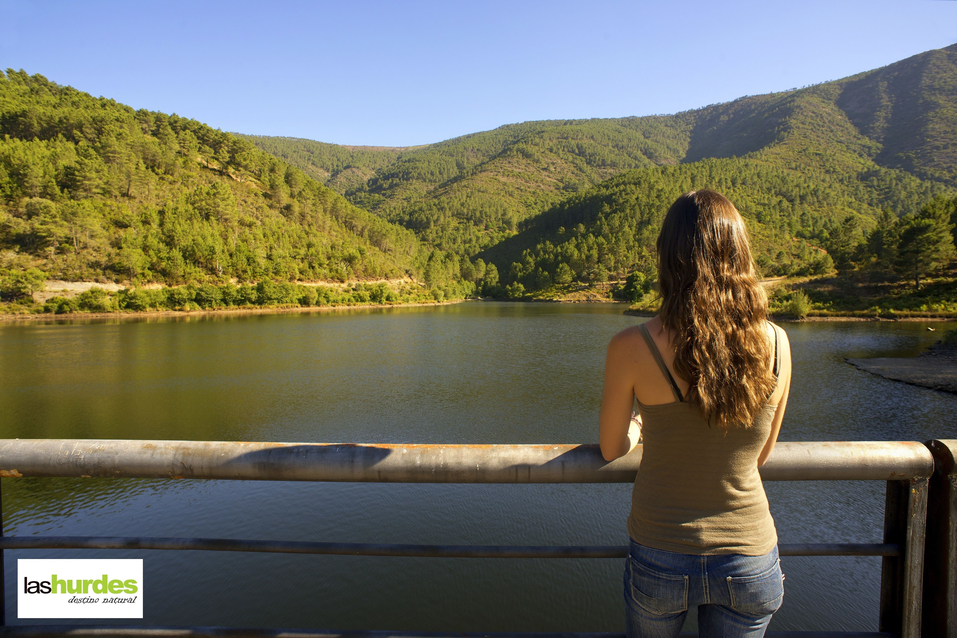 Une femme se tient près d’une balustrade, d’où elle a une vue sur un lac, avec des collines boisées en arrière-plan.