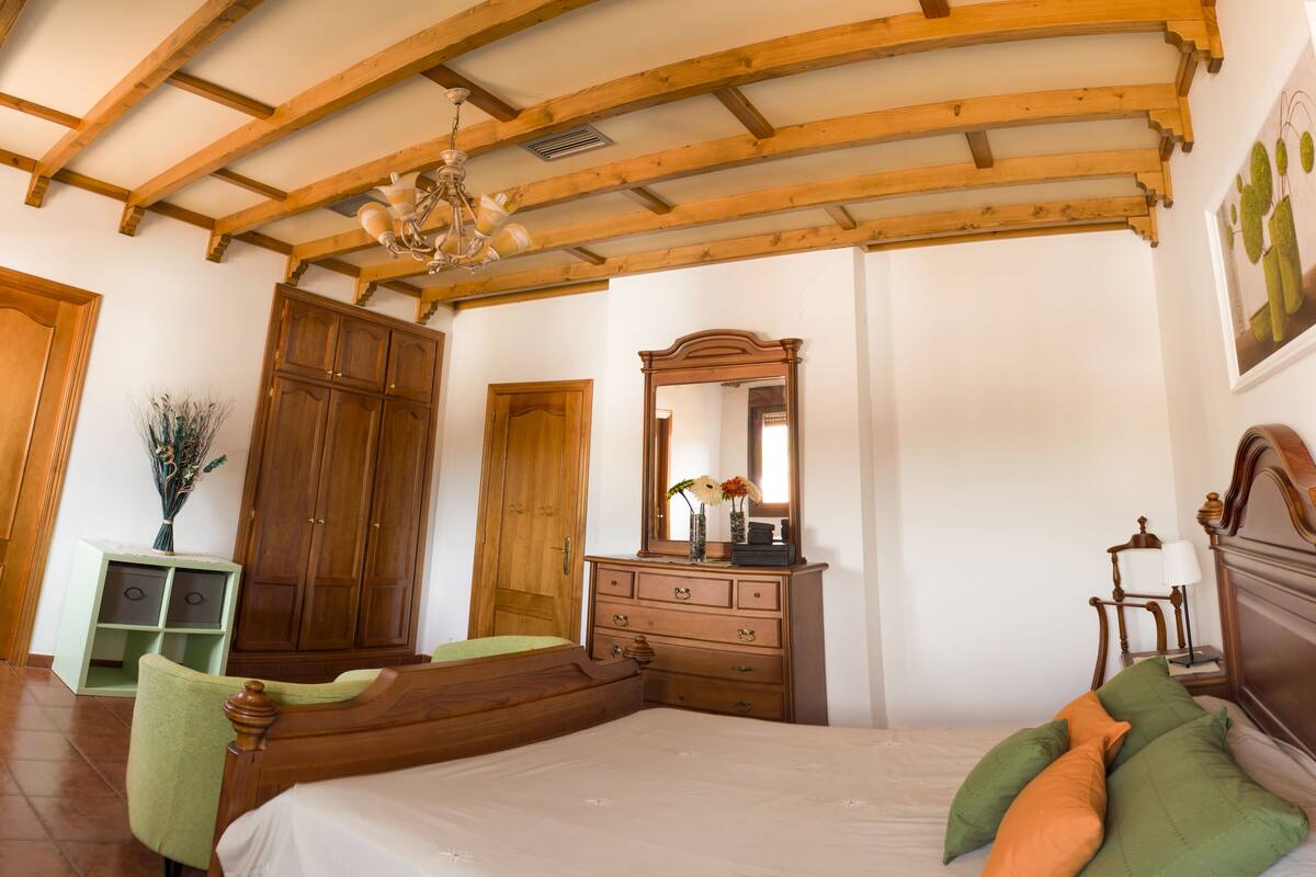 Une chambre avec un plafond en bois, un lit avec des coussins verts et orange, une commode avec un miroir et une chaise verte.