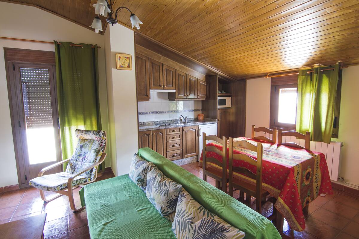 Une pièce chaleureuse avec un plafond en bois, une table à manger avec des chaises, un canapé, un fauteuil, un coin cuisine avec des placards et un four à micro-ondes, et une fenêtre avec des rideaux.