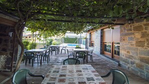 Een terras buiten met stenen muren, tafels, stoelen en een pergola begroeid met klimplanten.