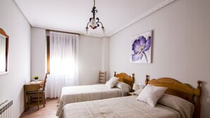 Een slaapkamer met twee bedden, een houten bureau, een stoel, een raam met gordijnen en een schilderij aan de muur.