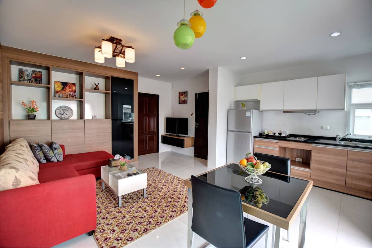 A modern living room with a red sofa, a glass coffee table, and a dining area with a black chair and a bowl of fruit.