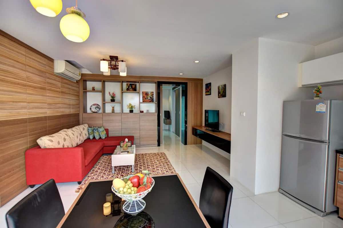 A modern living room with a red sofa, dining table with chairs, and a fruit bowl. There is a kitchen area with a refrigerator and a TV.