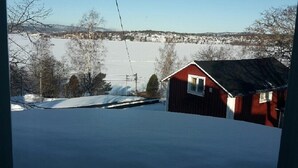 Une cabane rouge avec une fenêtre blanche, entourée de terre et d’arbres recouverts de neige, dominant un paysage enneigé.