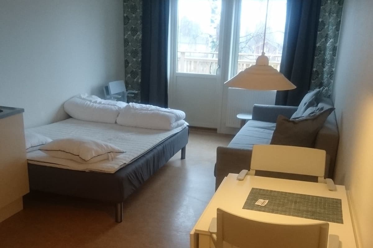 Une chambre d’hôtel de taille réduite, comprenant un lit simple, un petit bureau avec une chaise, une table et une lampe suspendue.