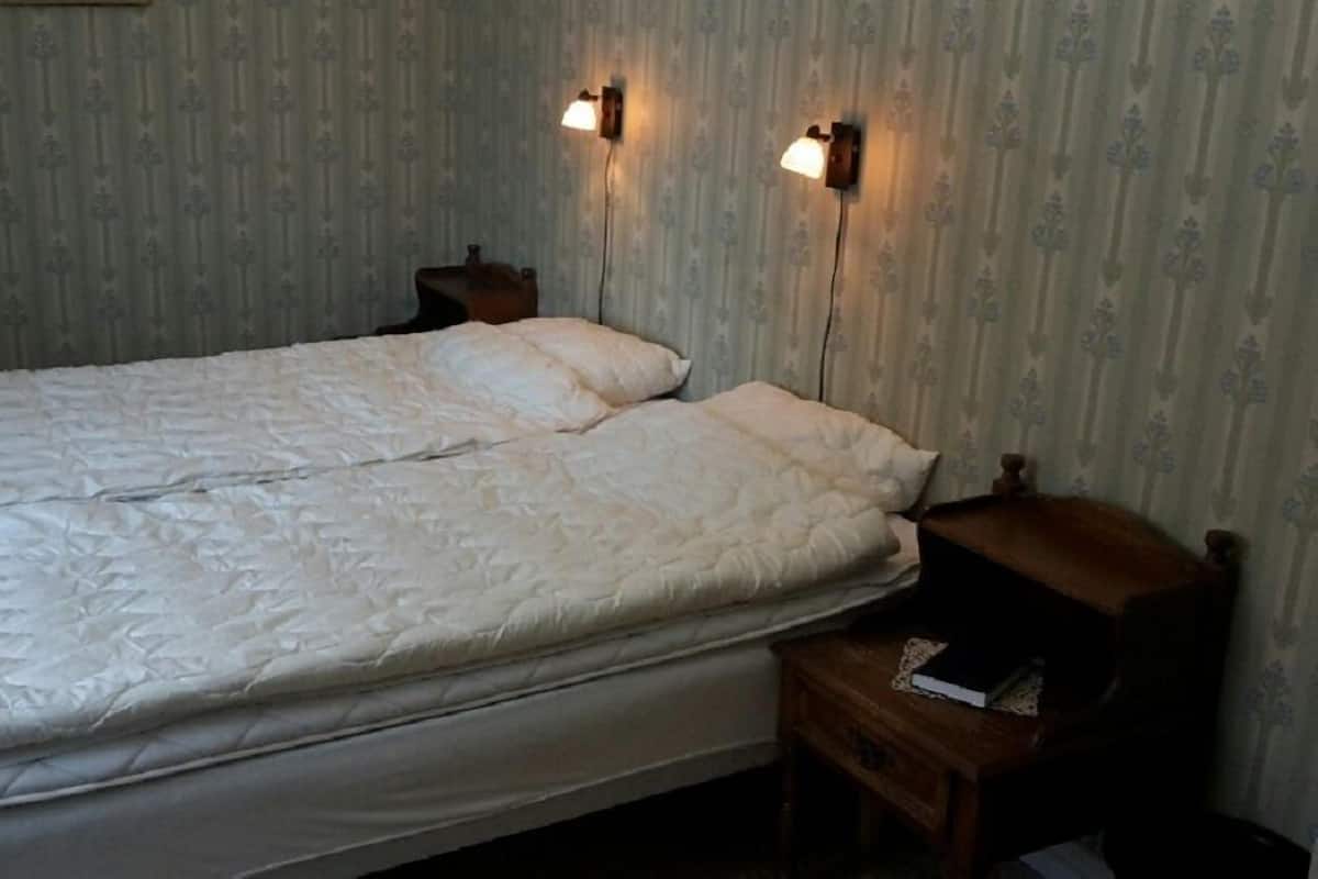 Un lit simple avec une couette blanche, dans une pièce dont le papier peint est à motifs et qui est équipée de deux lampes fixées au mur.