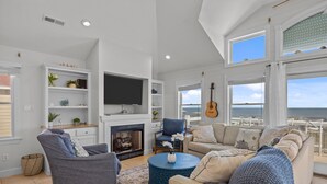 A spacious living room with a fireplace, a large TV, and a view of the ocean through large windows. - Atlantic Watch: Oceanfront | 14 bedrooms | Private Walkway | Heated Private Pool (Kill Devil Hills)