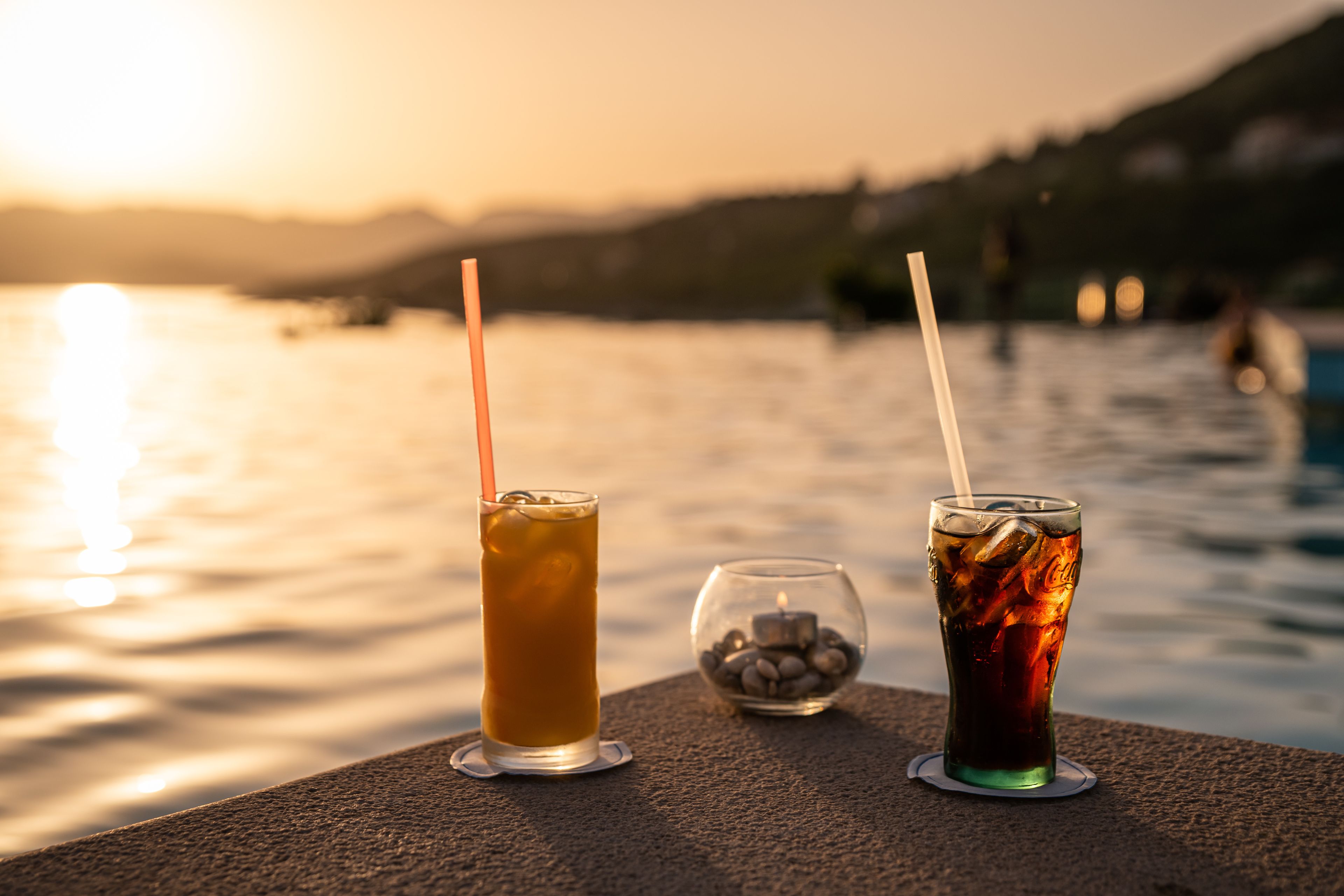 Two glasses of iced beverages on a poolside table with a candle and a glass of stones.