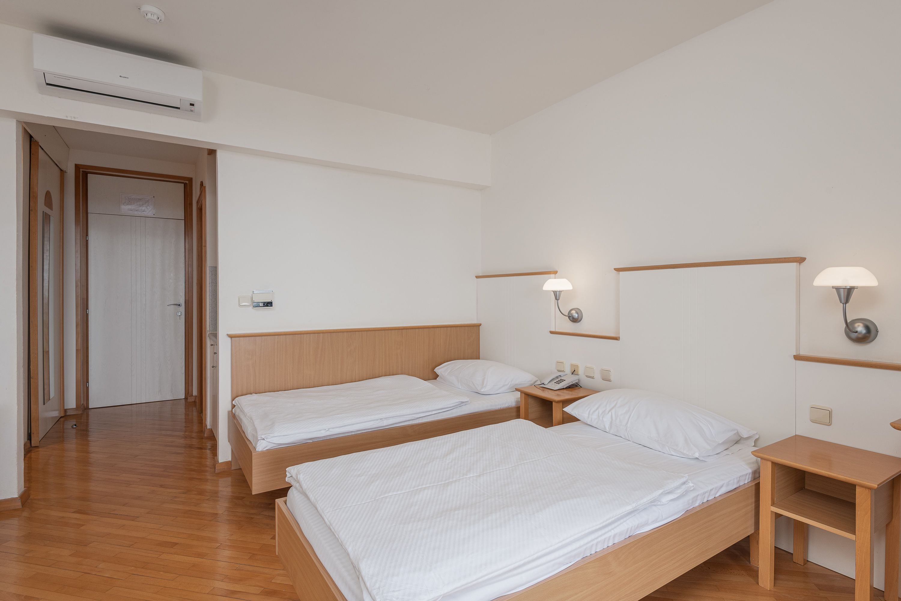 A hotel room with two single beds, wooden headboards, bedside tables, a nightstand with a lamp, and an air conditioning unit.