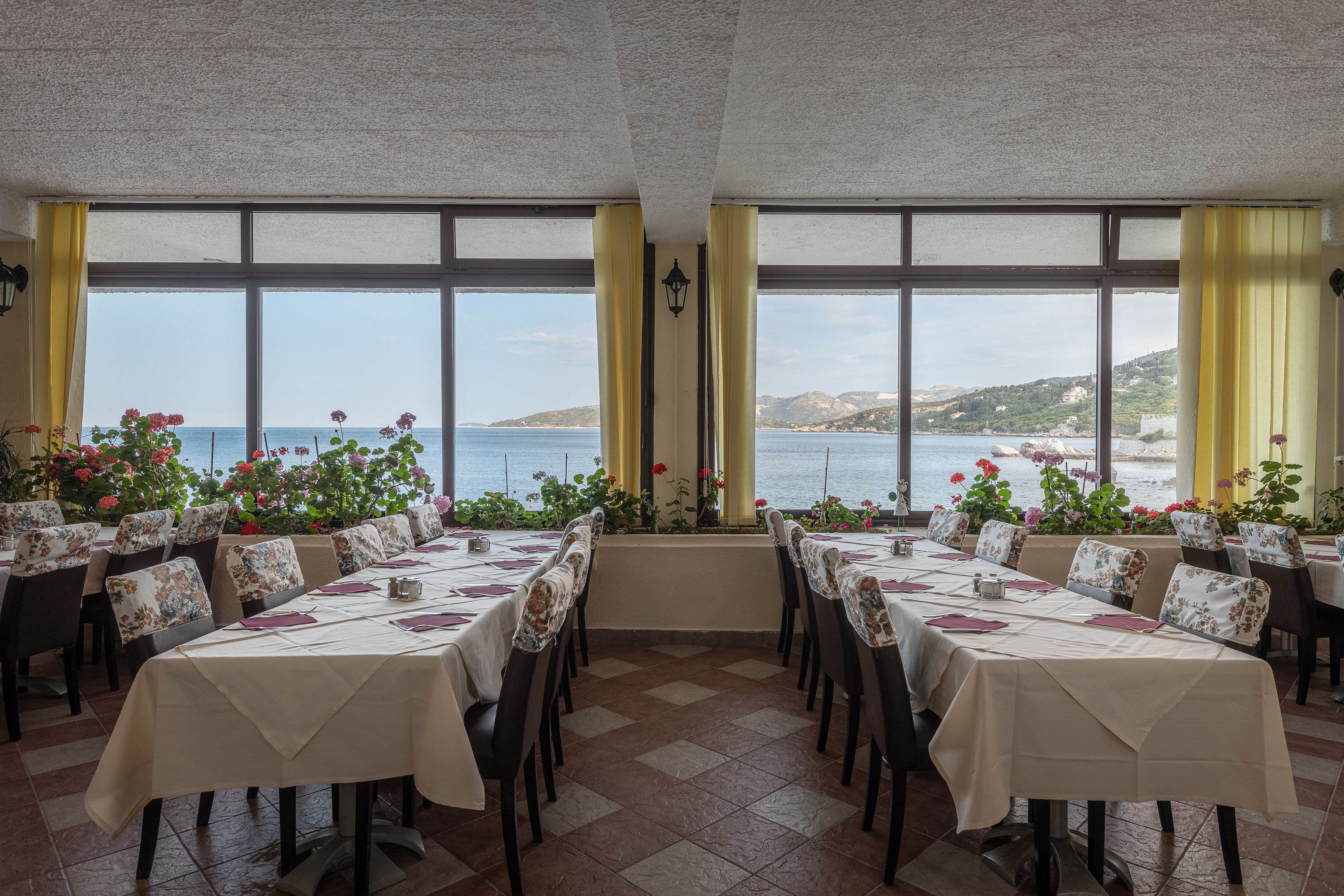 A restaurant with tables set for dining, large windows with a view of the sea, and potted plants on the windowsill.