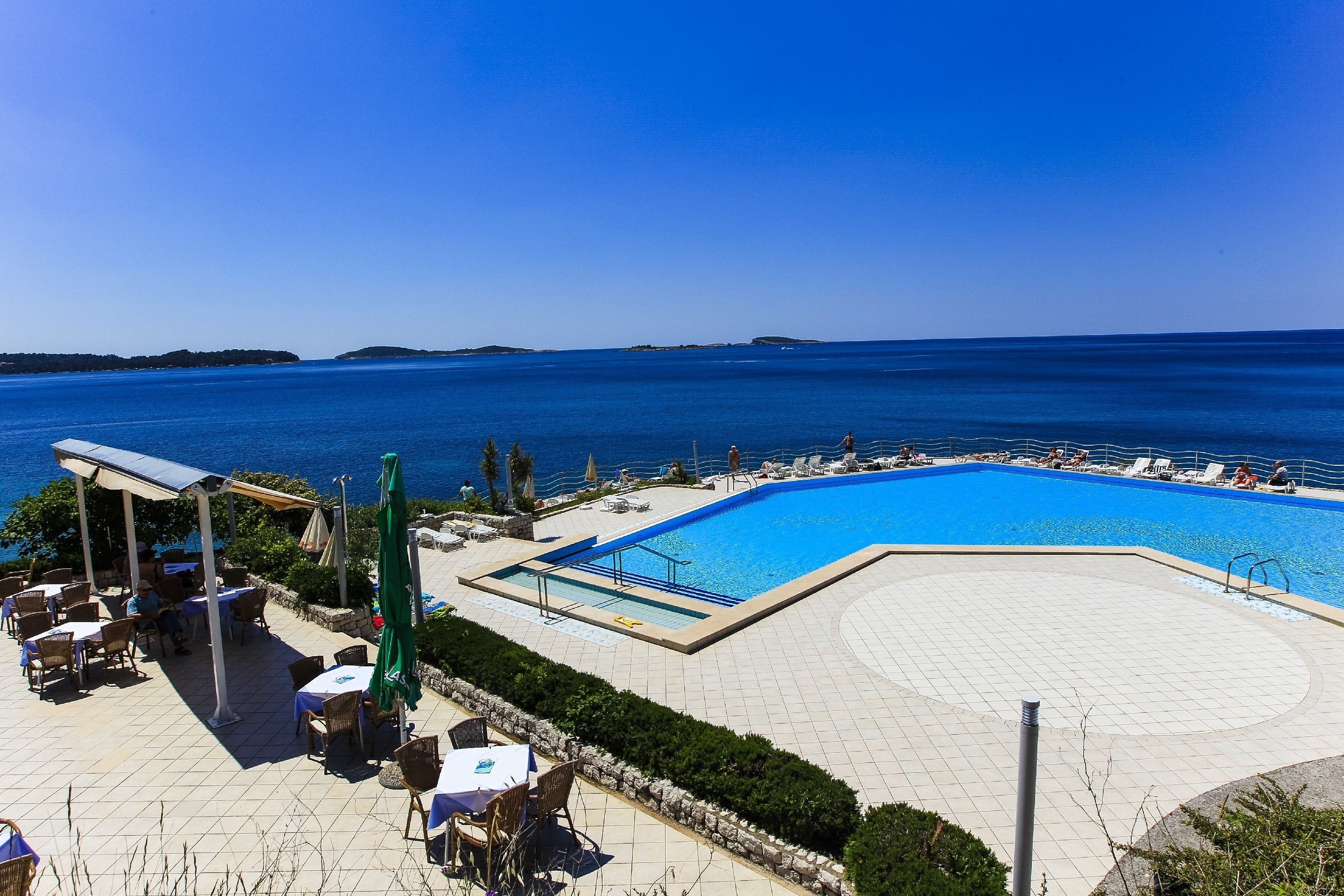 A poolside area with lounge chairs and tables, a pool, and a view of the sea.