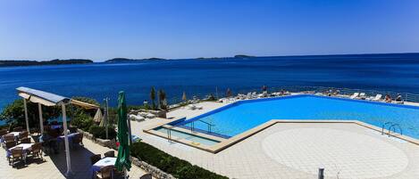A poolside area with lounge chairs and tables, a pool, and a view of the sea.