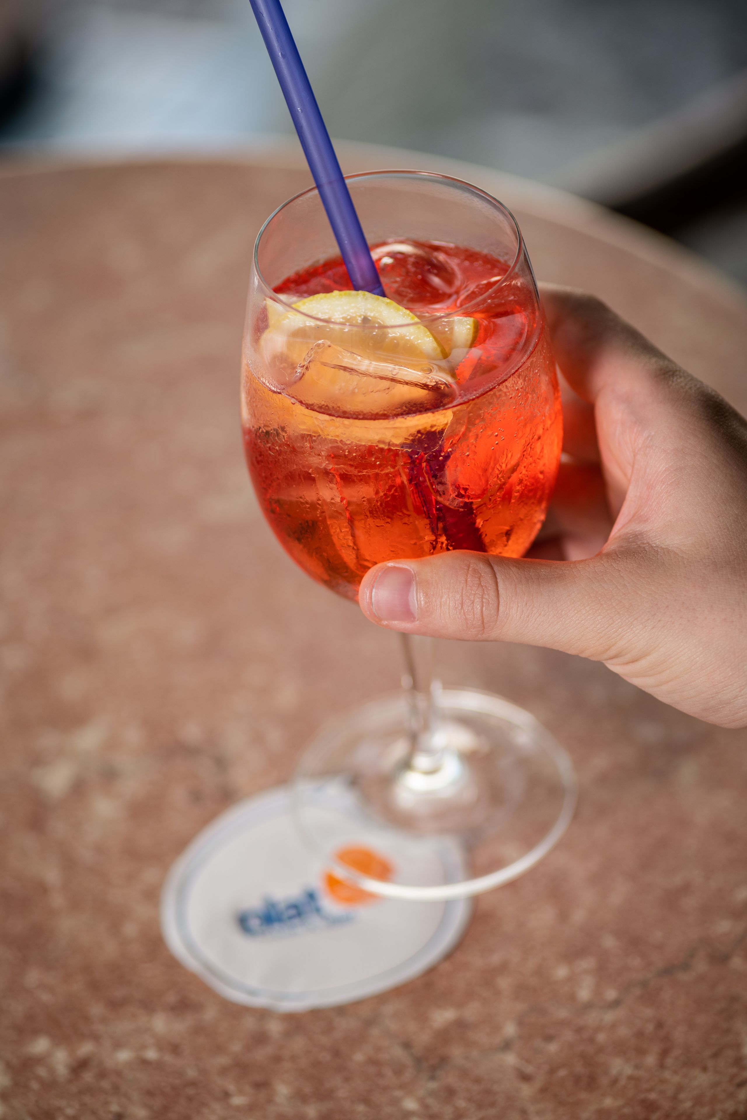 Une main tenant un verre avec une paille, contenant une boisson rouge et des tranches de fruits, et un dessous de verre portant un logo en dessous.