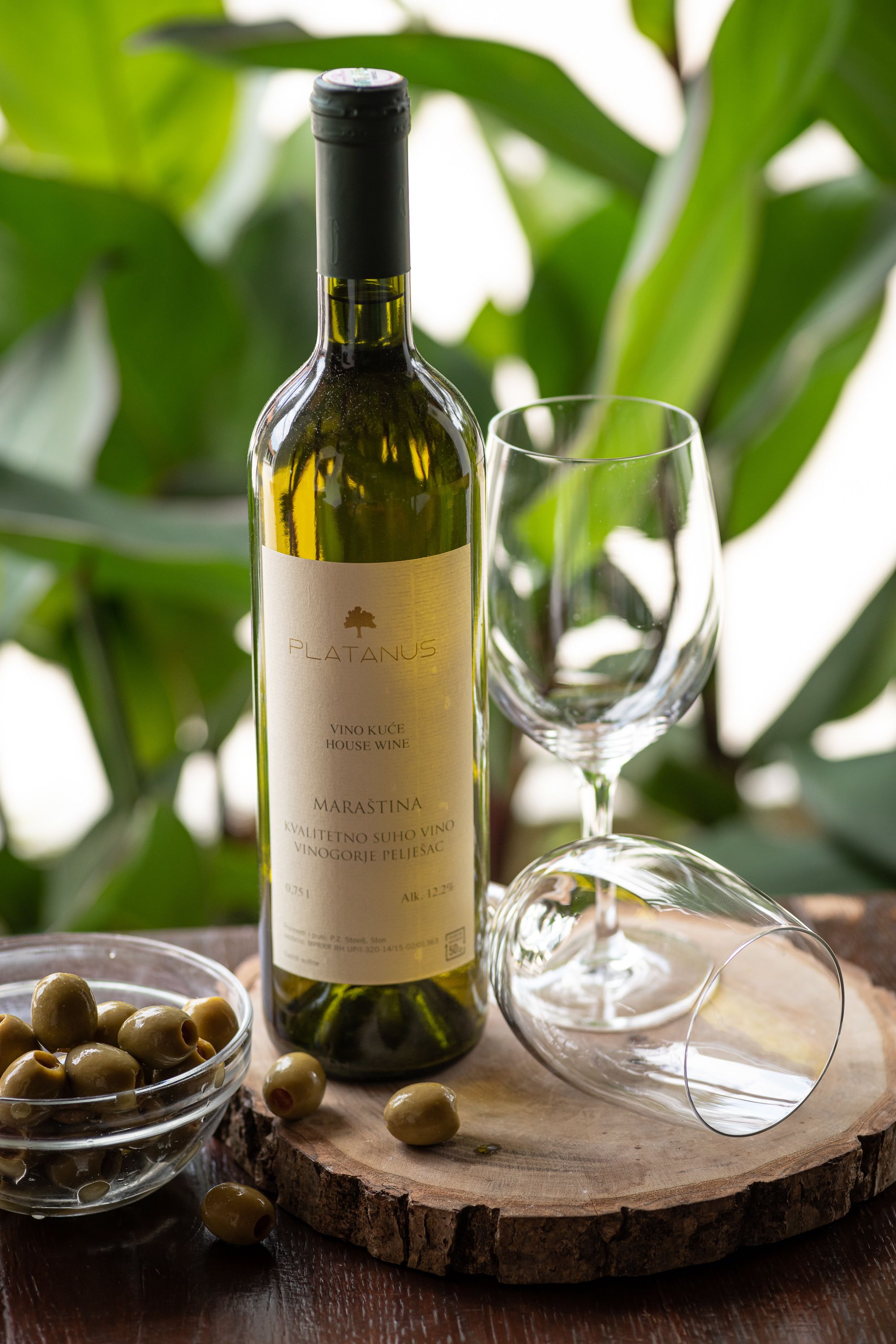 Una botella de vino Platanus Marastina, una copa y un cuenco de aceitunas sobre una superficie de madera.