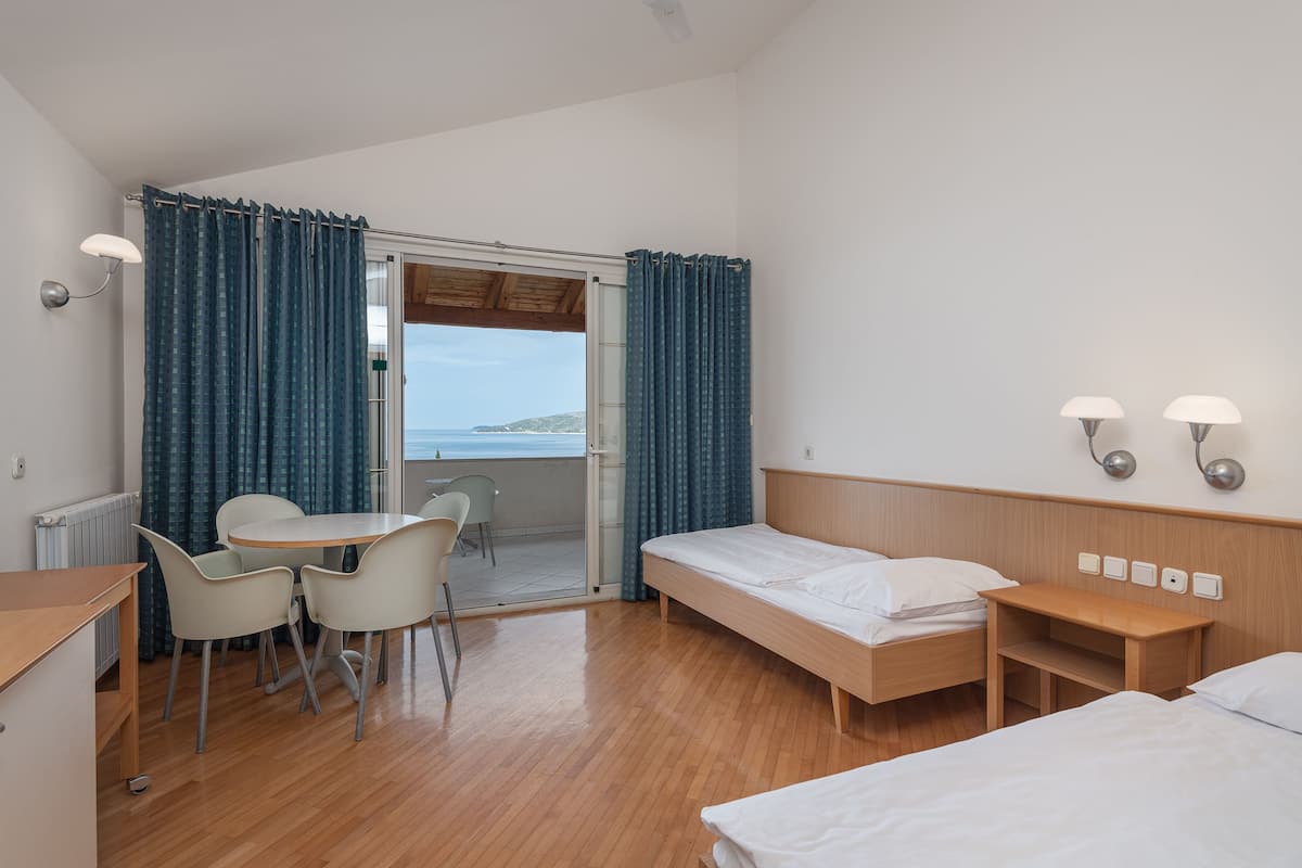 Une chambre d’hôtel avec un lit, un bureau, des chaises, une petite table et une vue sur la mer, visible par une porte ouverte.