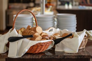 A buffet setup with bread and rolls in baskets, accompanied by serving utensils, and stacks of plates in the background.