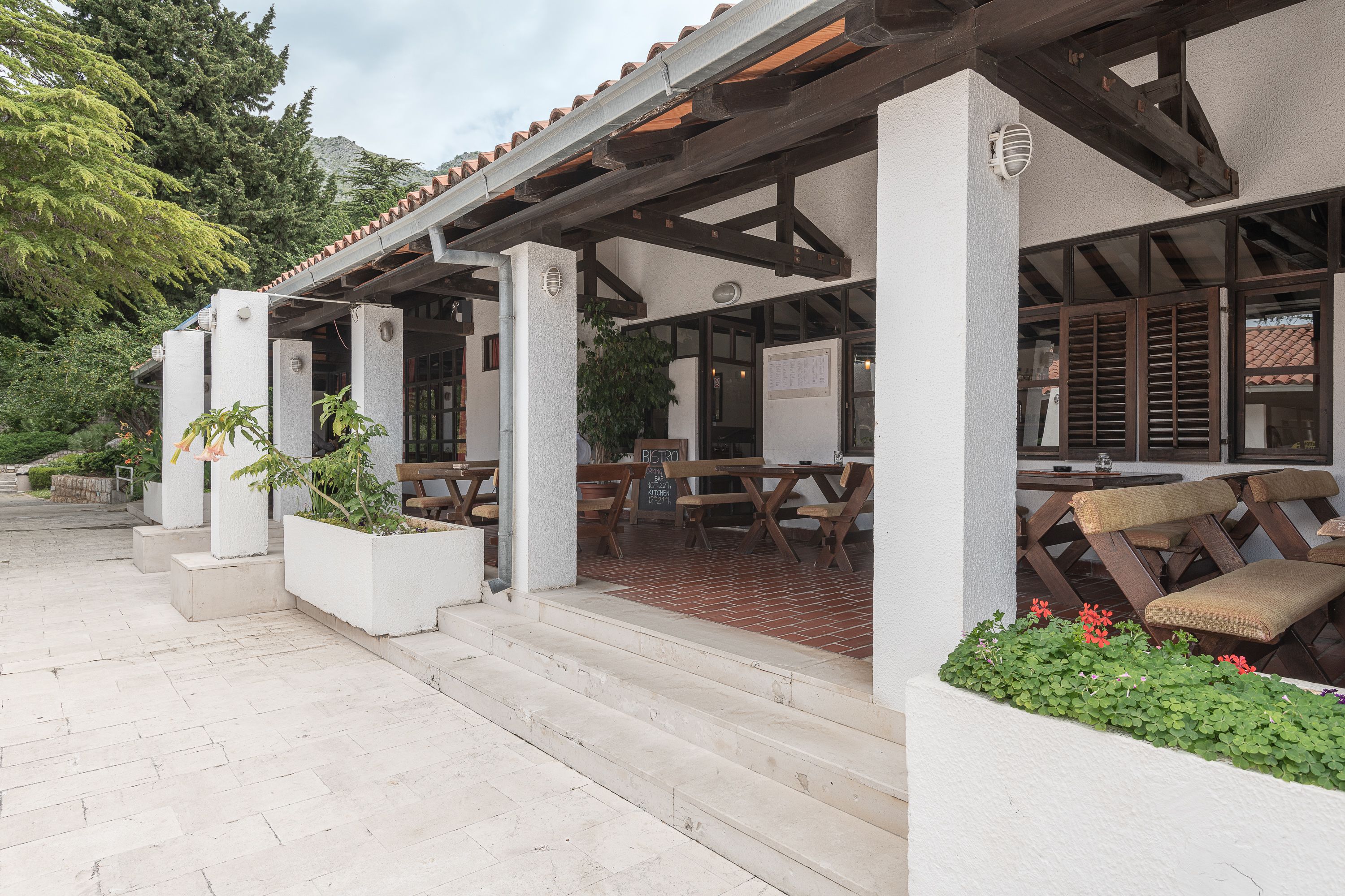 A covered outdoor seating area with tables and benches, surrounded by white walls and a tiled roof.