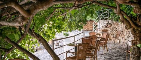 Une terrasse extérieure avec des chaises et des tables en osier, entourée de murs en pierre et d’un escalier en pierre.