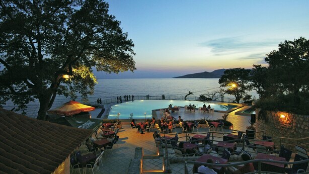 An outdoor pool area with seating and tables, surrounded by trees and overlooking a body of water.