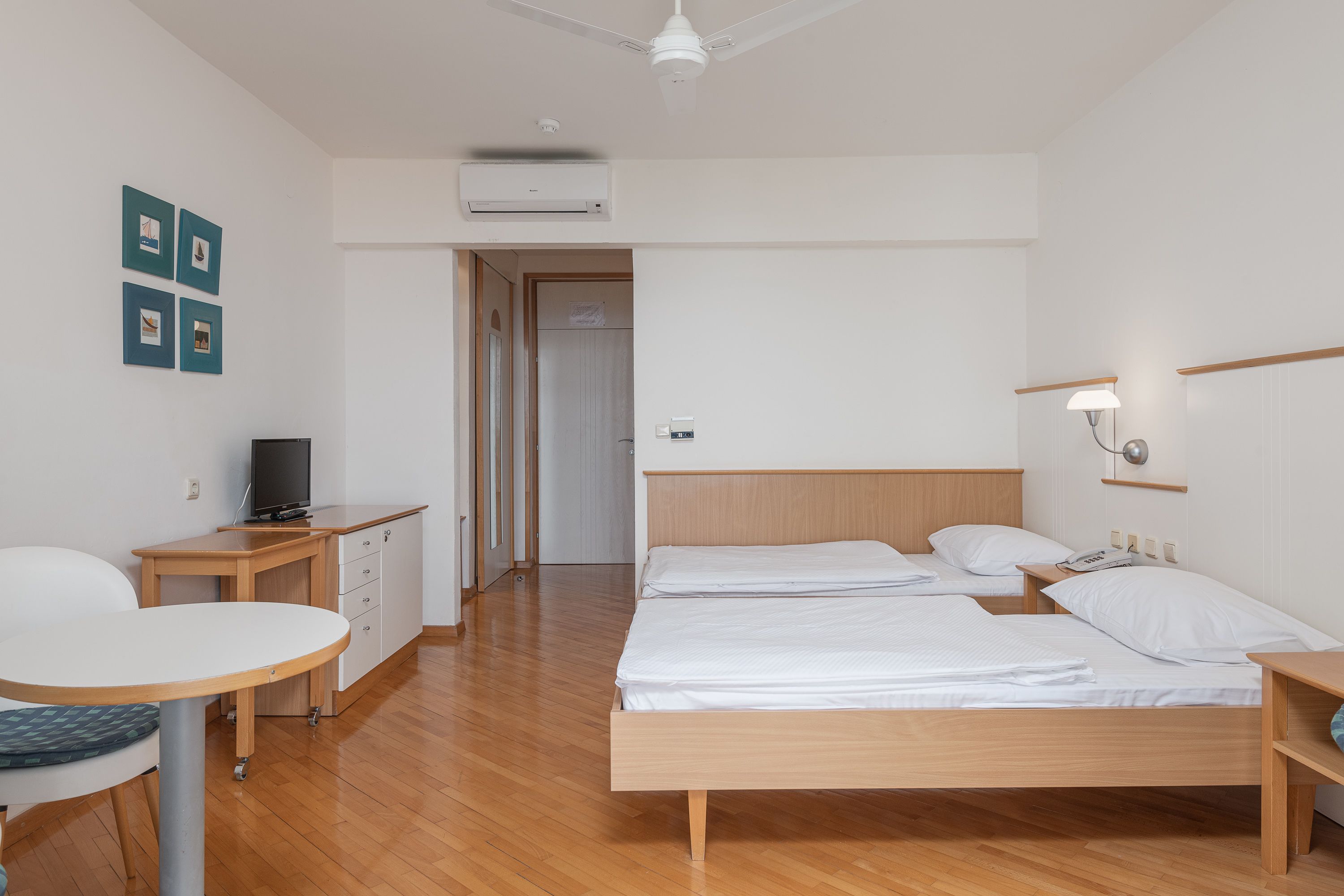 Een hotelkamer met een houten vloer, een bed met witte lakens, een bureau met een televisie en een klein tafeltje met een stoel.