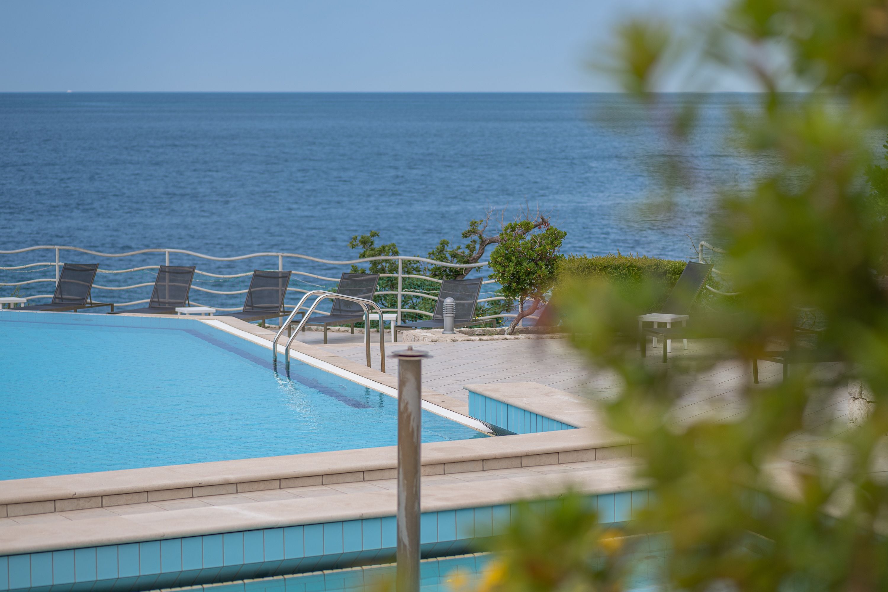 Une piscine avec vue sur l’océan, entourée de chaises longues et d’un petit jardin.