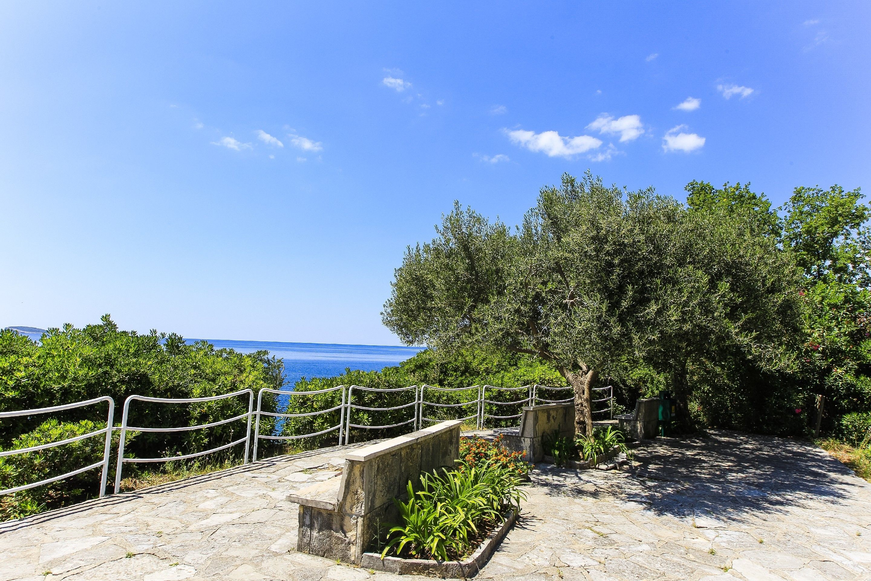A stone pathway leading to a scenic overlook with a metal railing, surrounded by greenery and overlooking a body of water.