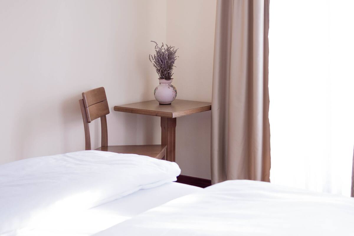 A hotel room with a bed, a wooden chair, a small table with a vase of flowers, and a window with curtains.