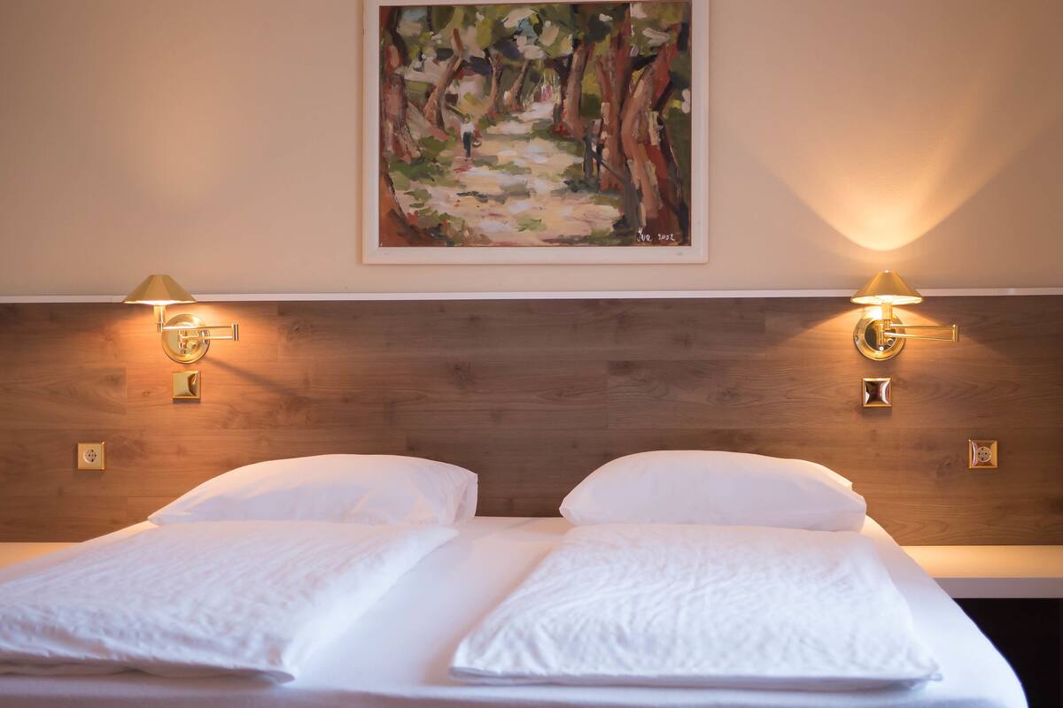 A double bed with white linens, two wall-mounted lamps, and a painting above the bed.