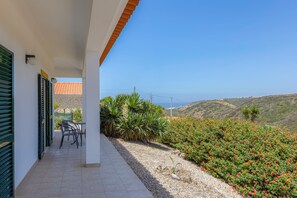 A balcony with a table and chairs, a view of the sea, and a garden with colorful flowers.