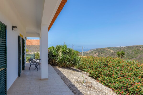 A balcony with a table and chairs, a view of the sea, and a garden with colorful flowers.