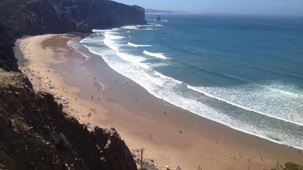 A coastal beach with a rocky cliff, clear blue sky, and calm sea.