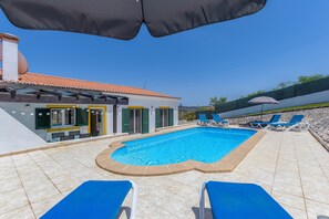 Une piscine entourée de chaises longues bleues, une terrasse carrelée et un bâtiment doté d’un toit et de volets également carrelés.