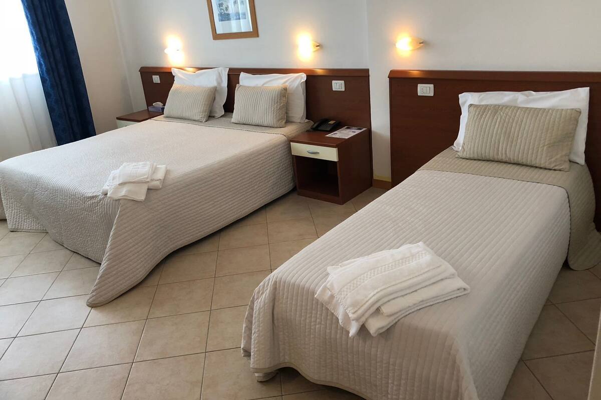 Habitación de hotel con dos camas individuales, cada una con una almohada y una toalla doblada. Se observa un mesita de noche con un teléfono y una pequeña mesa con una lámpara.