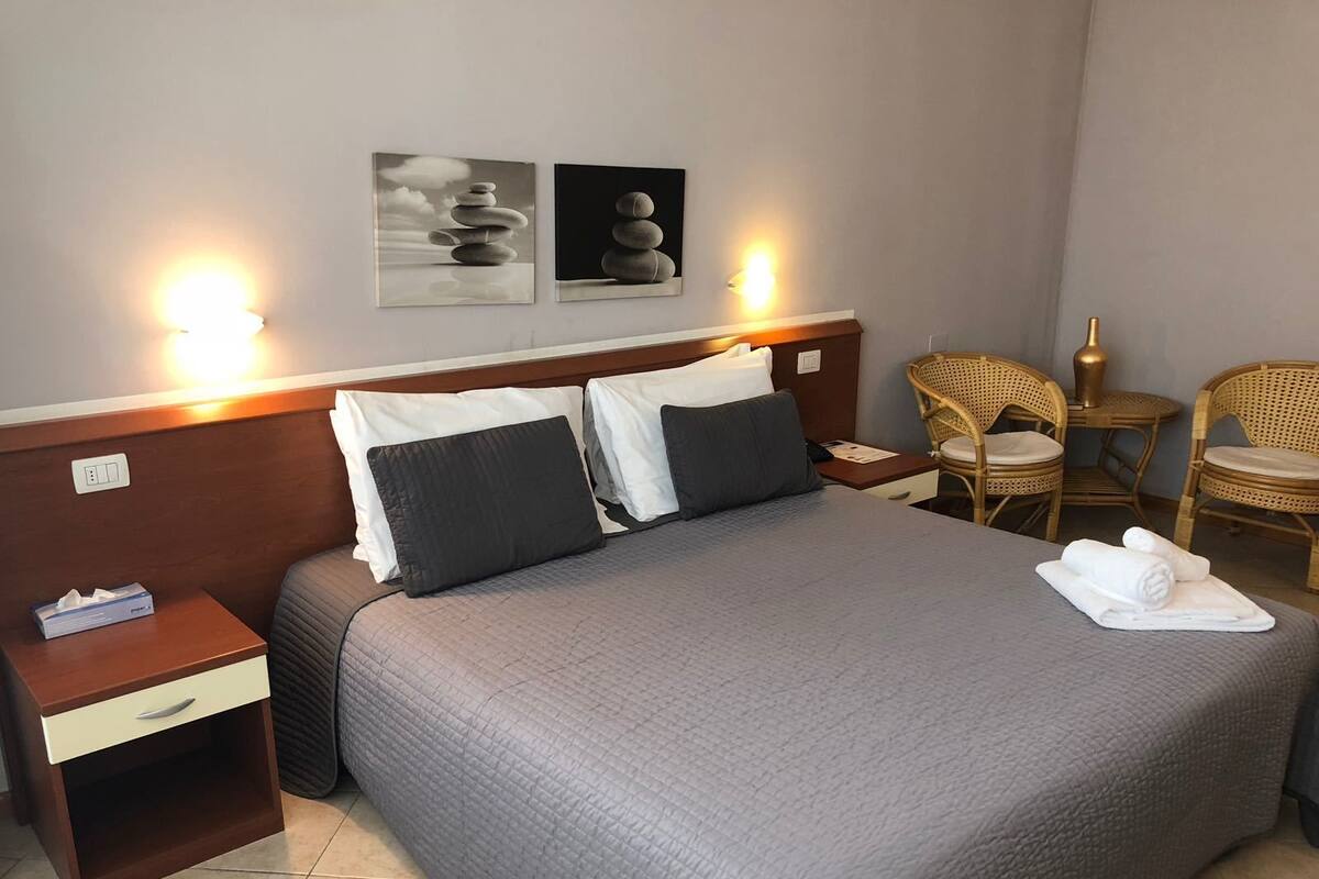 Una cama bien hecha con colcha gris, dos mesitas de noche con lámparas, una silla y dos obras de arte colgadas en la pared.