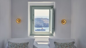 Une chambre à coucher avec un lit, deux coussins à motifs, une fenêtre donnant sur un navire et des montagnes, et deux appliques murales.