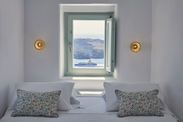 Un dormitorio con una cama, dos almohadones estampados, un ventanal con vista a un barco y montañas, y dos apliques de pared.