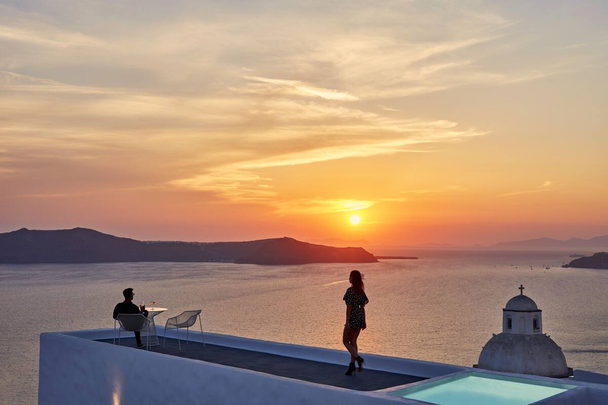 A sunset view from a rooftop terrace with a pool, two chairs, and a small building with a dome.