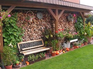 A wooden bench under a pergola with a stone wall behind it, surrounded by potted plants and a neatly trimmed lawn. - Ferienwohnung Kerstin (Siggelkow)
