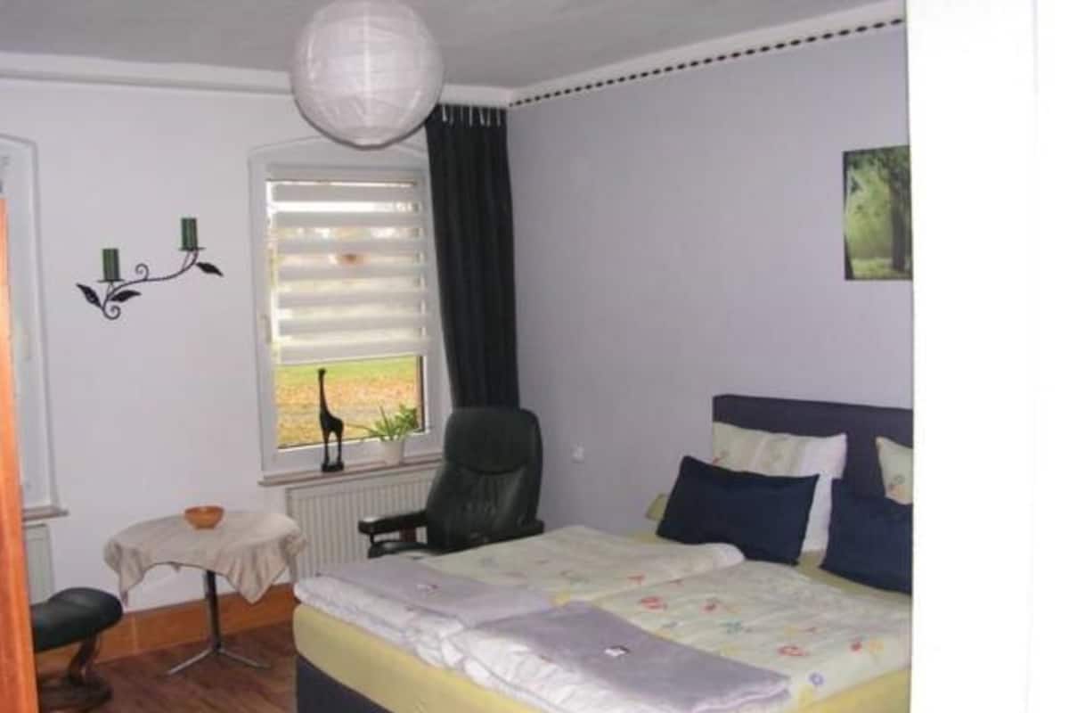 A bedroom with a bed, a chair, a small table, and a window with blinds.