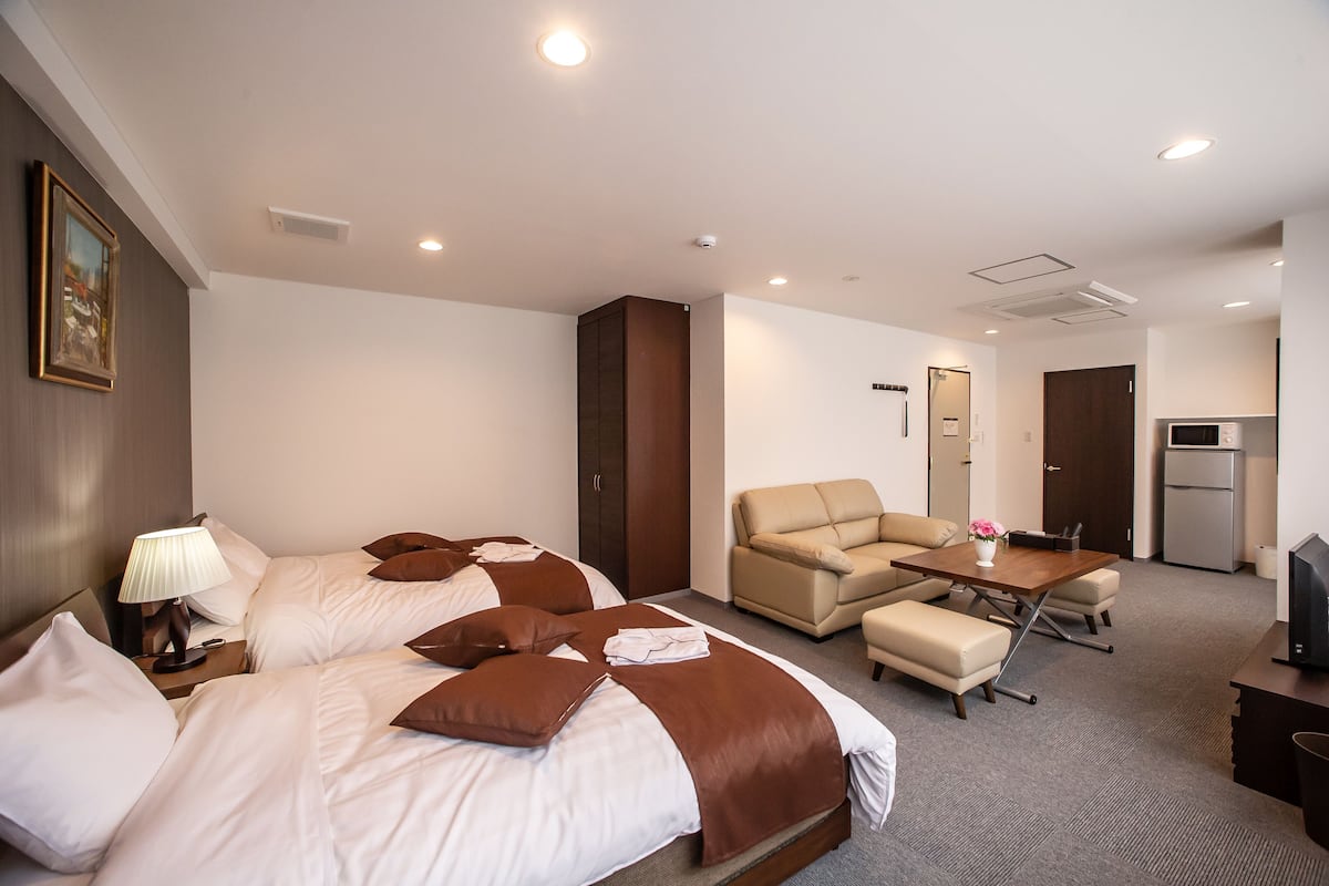 A hotel room with two beds, a sofa, a small table, a TV, and a kitchenette.